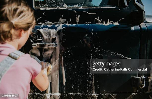 a little girl uses a yellow sponge to wash a blue car, the soap creates a lather that runs down the side of the car - hubcap stock pictures, royalty-free photos & images