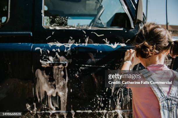 a little girl uses a yellow sponge to wash a blue car, the soap creates a lather that runs down the side of the car - hubcap stock pictures, royalty-free photos & images