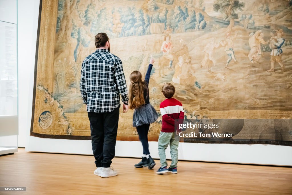 Young Girl Pointing Up Towards Large Painting While Exploring Museum With Family