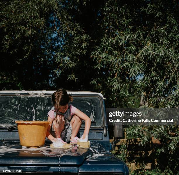 a little girl attempts to be helpful, climbing onto the bonnet of a vintage 4x4 and washing it with sponges - car detailing stock pictures, royalty-free photos & images