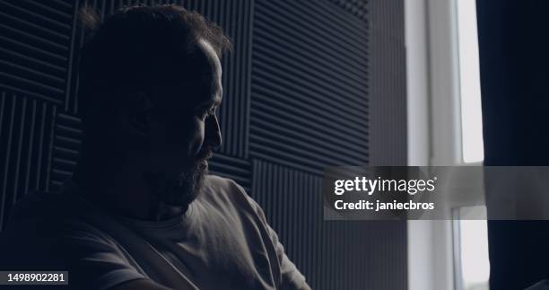 depressed man alone in a dark room. sitting by the window and looking out. rubbing bridge of nose - head in hands silhouette stock pictures, royalty-free photos & images