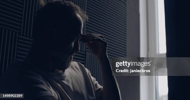 depressed man alone in a dark room. sitting by the window and looking out with worry. rubbing bridge of nose - head in hands silhouette stock pictures, royalty-free photos & images