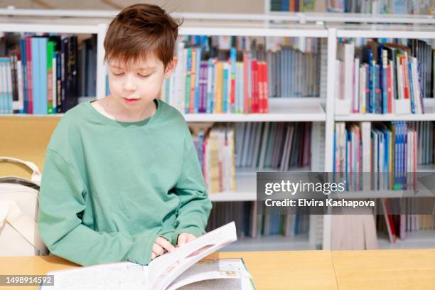 a shaggy boy of 10 years old in a green t-shirt sits in the library against the background of bookshelves and reads a book - 10 11 years boy stock-fotos und bilder