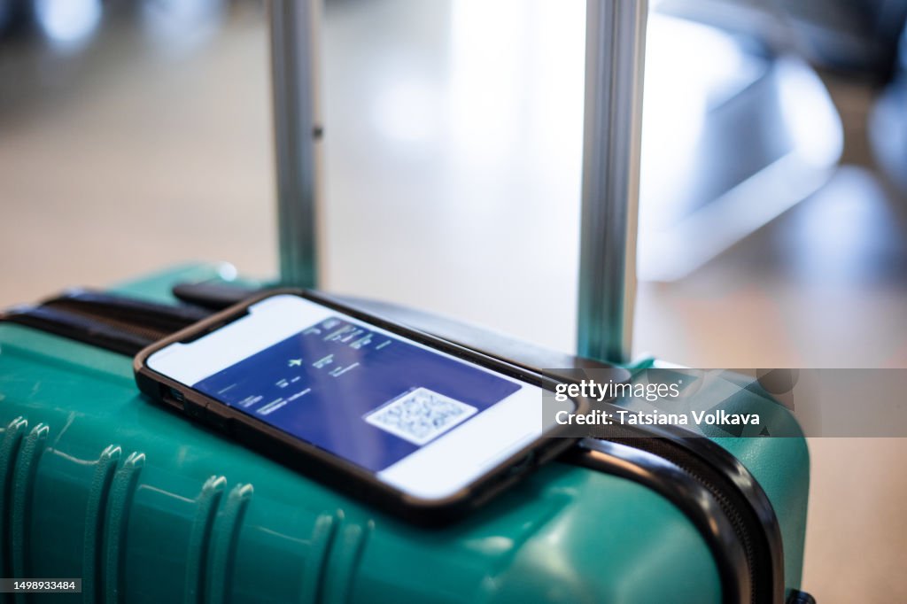 Close-up image of mobile phone screen displaying plane ticket lying on top of rolling suitcase