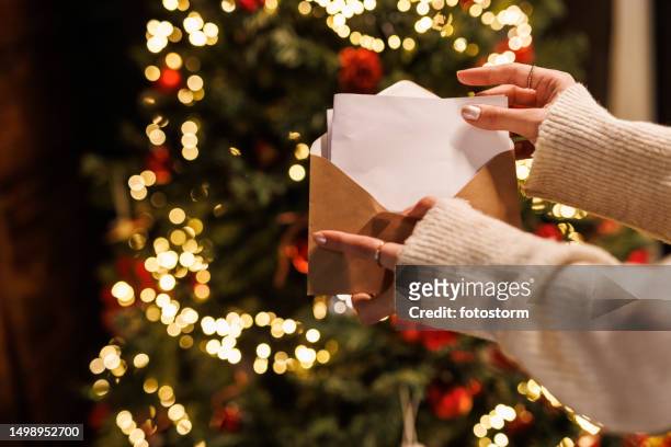 young woman standing by the glistening christmas tree and opening a letter - envelope stockfoto's en -beelden