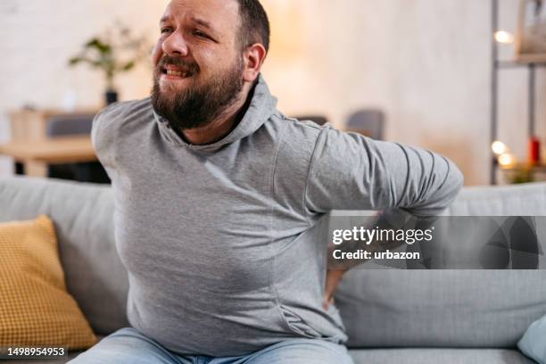 stressed man suffering back pain while sitting on the sofa - 下背部痛 個照片及圖片檔