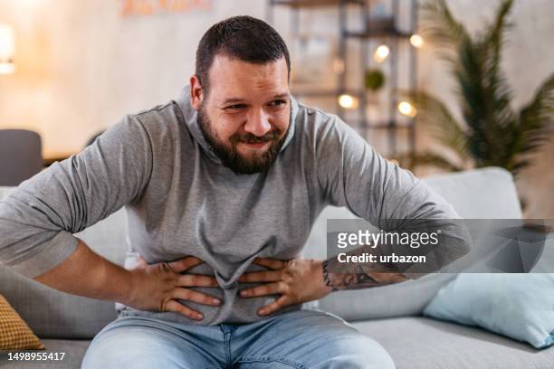 man with severe stomach pain sitting at home. - fat guy belly stock pictures, royalty-free photos & images