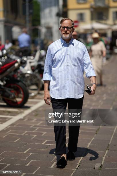 Bruce Pask wears black glasses, a white t-shirt, a pale blue and blue striped print pattern shirt, black suit pants, black velvet loafers , outside...