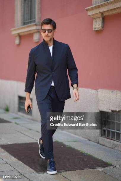 Johannes Huebl wears black sunglasses, a white t-shirt, a navy blue high neck blazer jacket, matching navy blue suit pants, navy blue shiny leather...