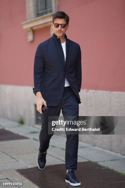 Johannes Huebl wears black sunglasses, a white t-shirt, a navy blue high neck blazer jacket, matching navy blue suit pants, navy blue shiny leather...