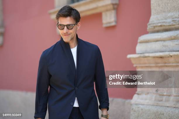 Johannes Huebl wears black sunglasses, a white t-shirt, a navy blue high neck blazer jacket, matching navy blue suit pants, a khaki shiny leather...