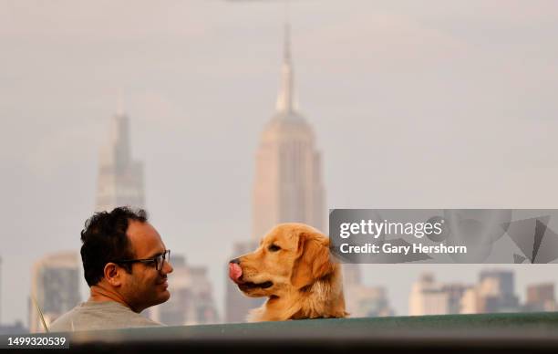 Man sits with his golden retriever dog in front of the skyline of midtown Manhattan and the Empire State Building as the sun sets in New York City on...