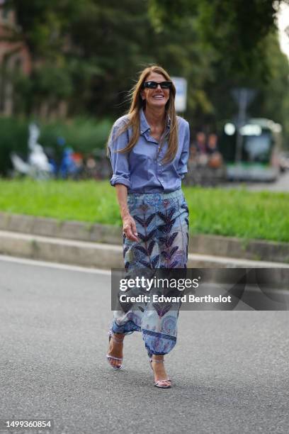 Anna Dello Russo wears black sunglasses, diamonds small earrings, a blue cotton shirt, a pale blue with purple / navy blue print pattern and...