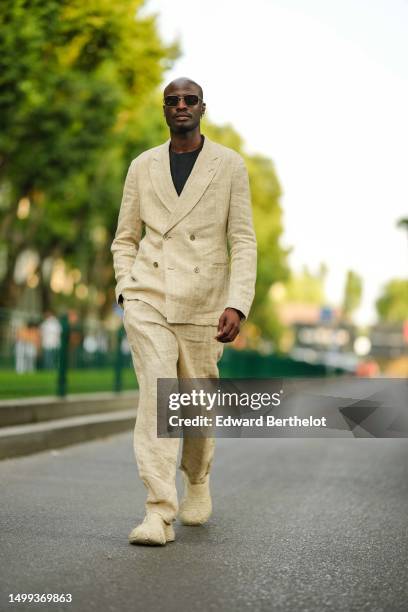 Guest wears black sunglasses, silver earrings, a black t-shirt, a beige linen blazer jacket, matching beige linen suit pants, beige embossed pattern...