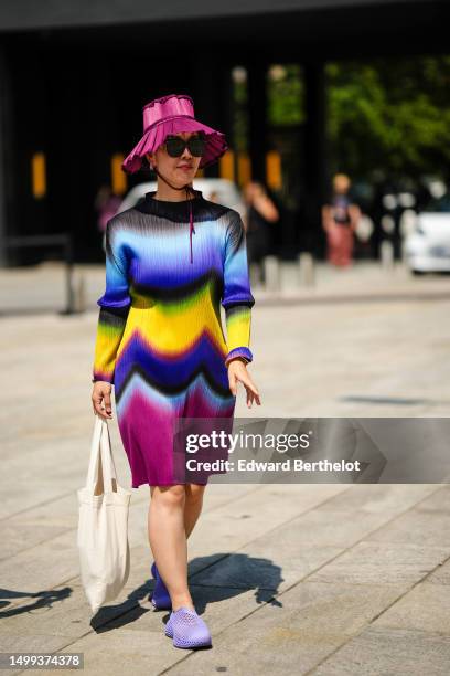 Guest wears a purple and pink linen ruffled bob hat, black large sunglasses, a blue / yellow / purple / wavy print pattern long sleeves knees dress,...