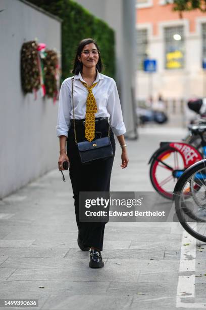 Guest wears glasses, silver earrings, a white shirt, a yellow with black flower print pattern tie, a black shiny leather crossbody bag, a black shiny...