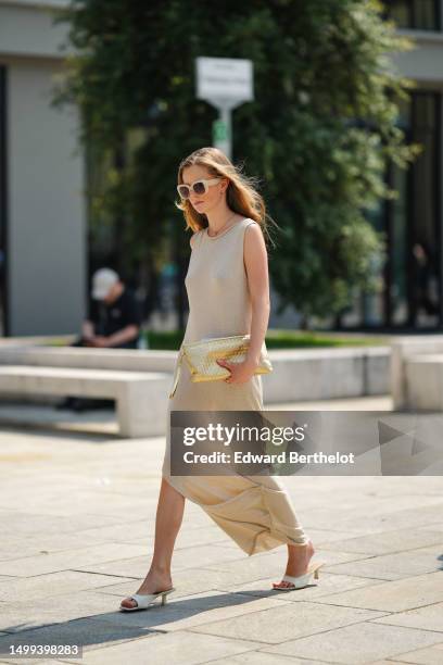 Guest wears beige plastic large sunglasses, micro gold earrings, a gold long necklace, a beige wool sleeveless long slit / split dress, white shiny...