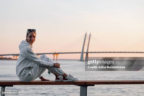 charismatic female solo traveler in white tracksuit sitting on shore of gulf of finland at sunset - ballustrade stockfoto's en -beelden