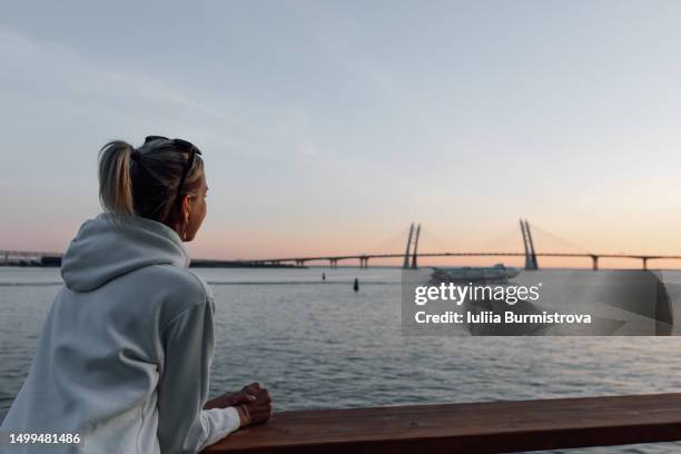 adventurous female traveler with ponytail standing on shore of gulf of finland viewing bolshoy obukhovsky bridge - balustrade foto e immagini stock