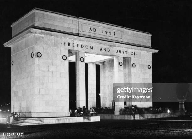 People sitting beneath the Black Star Gate, with the inscription 'AD 1957' above 'Freedom and Justice', floodlit by night, in Independence Square in...