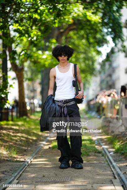 Guest wears black sunglasses, a white pearls necklace, a black shiny leather oversized kimono jacket, a black and white inscription print pattern...