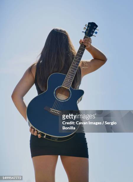 a teenage female guitarist holds an acoustic guitar to her back against a blue sky - custodia per chitarra foto e immagini stock
