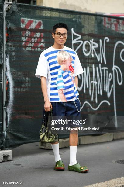 WeiRan Ji wears black glasses, a white with navy blue large striped print pattern and child print pattern t-shirt, navy blue denim large shorts, a...