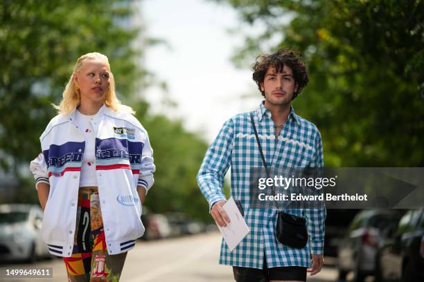 Almuneda Lapique wears gold large earrings, a white ribbed tank-top, a white with embroidered navy blue / red striped pattern oversized bomber coat,...