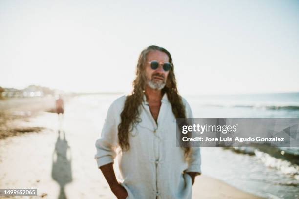 medium shot of mature man with long loose hair, wears a beard in sunglasses smiles and looks at camera on the beach in summer - medium length hair photos et images de collection