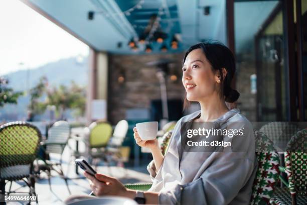 cheerful young asian woman using smartphone while sitting at a sidewalk cafe alone during free time, enjoying time on her own on a beautiful sunny day. lifestyle and technology. slow living. sustainable lifestyle - asian woman dream stock pictures, royalty-free photos & images