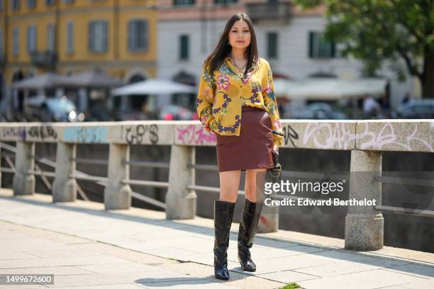 Guest wears gold earrings, a gold chain with lock pendant necklace, a yellow with multicolored flower print pattern silk shirt, a dark brown shiny...