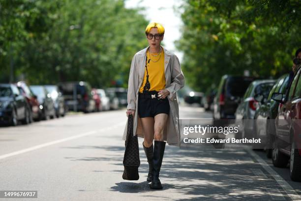 Guest wears black sunglasses, a yellow with black print pattern t-shirt, silver necklaces, a beige long coat, black nylon micro shorts, a black and...