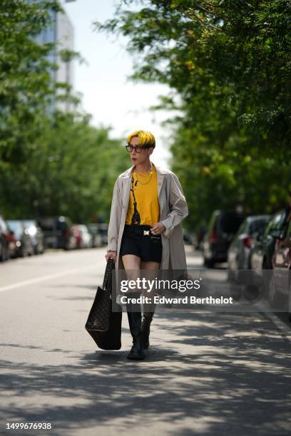 Guest wears black sunglasses, a yellow with black print pattern t-shirt, silver necklaces, a beige long coat, black nylon micro shorts, a black and...