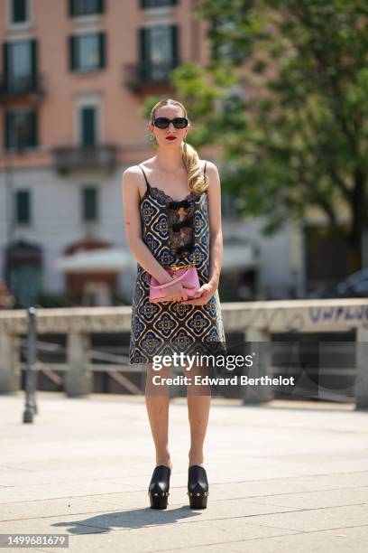 Guest wears black sunglasses, gold and green pendant earrings, a black lace print pattern V-neck / blue beige and brown print pattern tank-top knees...