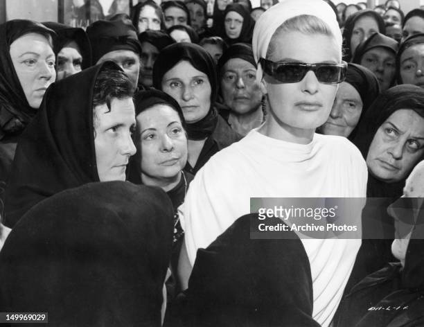 Melina Mercouri wearing sunglasses and surrounded by local women in a scene from the film 'Phaedra', 1962.