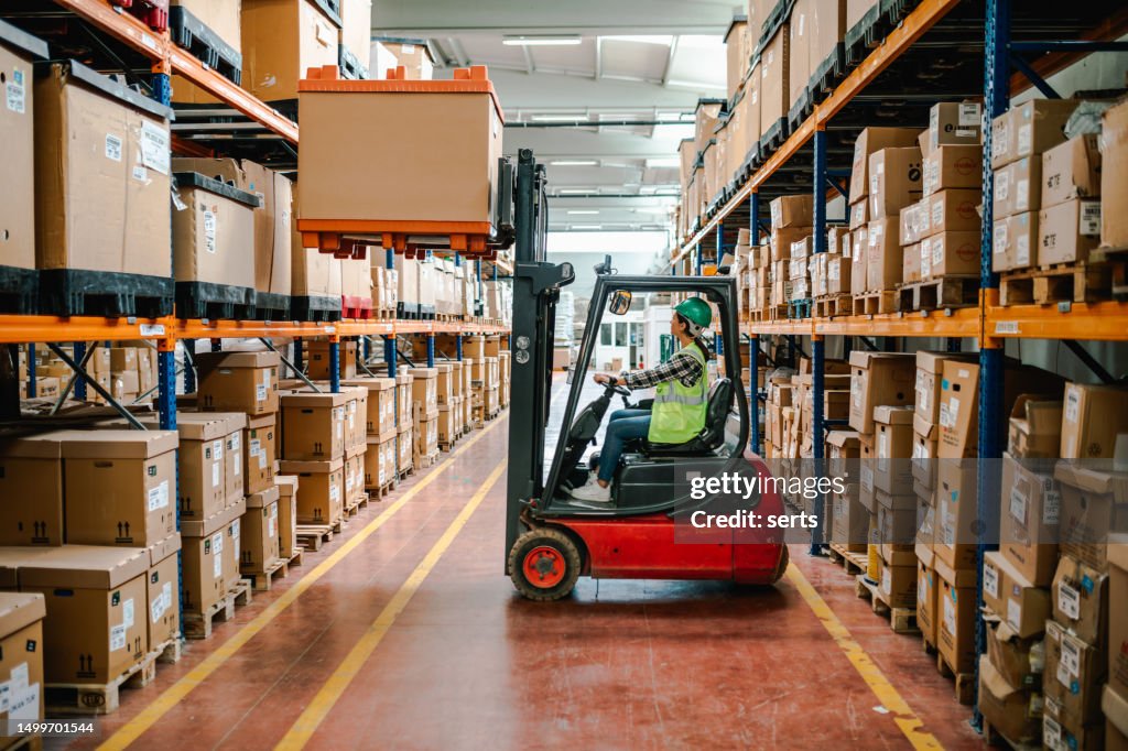 Efficient Warehouse Logistics: Young Woman Forklift Driver Moving Boxes onto Truck at Loading Dock in a Factory Warehouse