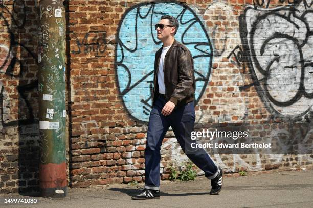 Guest wears black sunglasses, a white t-shirt, a dark brown faded leather zipper bomber coat, navy blue denim pants, black shiny leather and white...