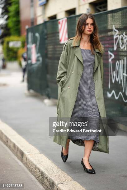 Guest wears a khaki long oversized coat, a white with black checkered print pattern oversized midi dress, black shiny leather pointed heels shoes ,...