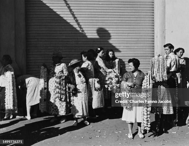 Group of Hawaiian people, each holding armfuls of leis on a street, standing before a metal shutter, Hawaii, circa 1955.