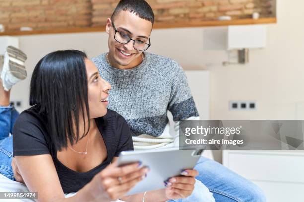 a young couple hugging and smiling while lying down and watching something on a tablet in a white room with wooden details. - private terrace balcony stock pictures, royalty-free photos & images