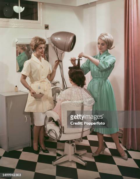Posed studio portrait of three female fashion models in a hair salon scene, the model on left wears a yellow nylon shortie overall and, on right, a...