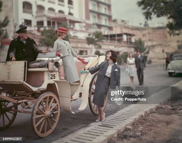 Posed vacation scene of two female fashion models posed on and beside a horse drawn carriage in a resort setting, the model on left wears a light...