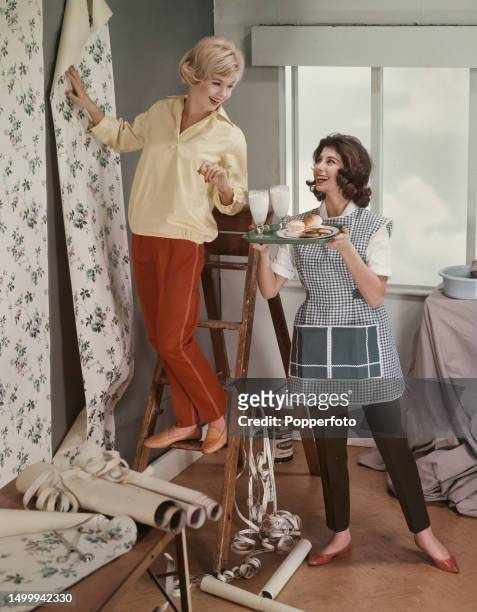 Posed studio portrait of two female fashion models in a home decorating scene, the model on left wears a yellow smock over a pair of scarlet...