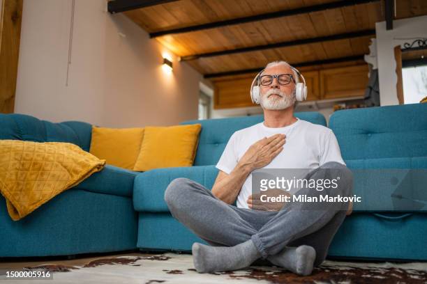 mature senior man sitting on floor practicing guided meditation at home, relaxing body and mind concept - groom stockfoto's en -beelden