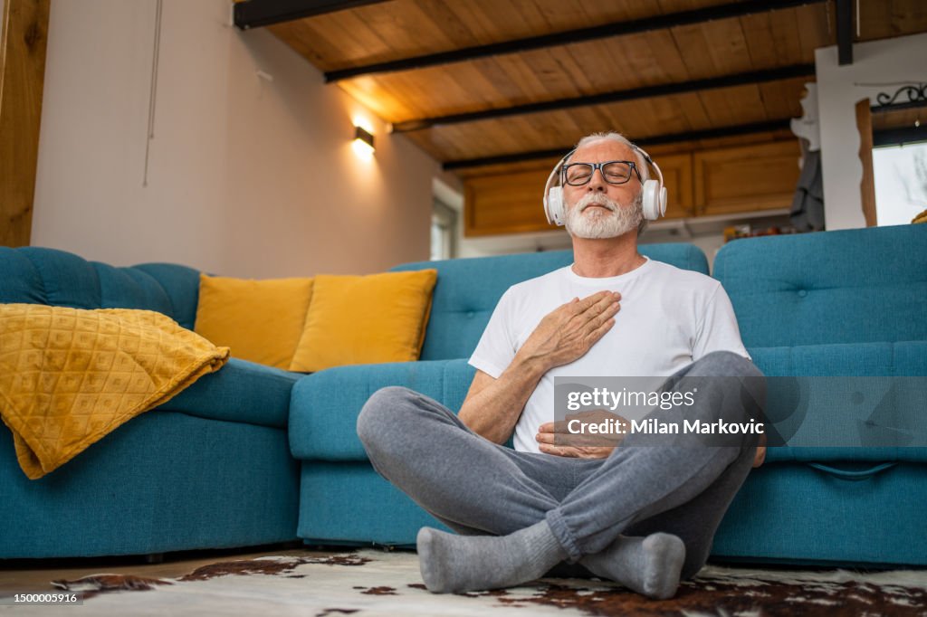 Mature senior man sitting on floor practicing guided meditation at home, relaxing body and mind concept