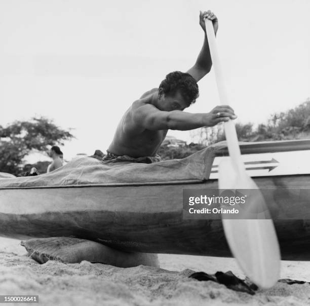 Man raises his oar as he sits in a Hawaiian canoe, which is raised off the sands of a beach on sacks, on the island of Oʻahu, Hawaii, circa 1955.