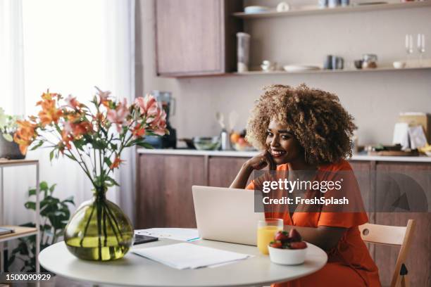 erfolgreiche afroamerikanische geschäftsfrau sitzt am küchentisch und liest einen bericht auf ihrem laptop - woman portrait kitchen laptop stock-fotos und bilder