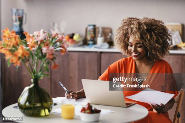 successful african american businesswoman sitting at the kitchen table, reading a report on her laptop - orange dress stock pictures, royalty-free photos & images