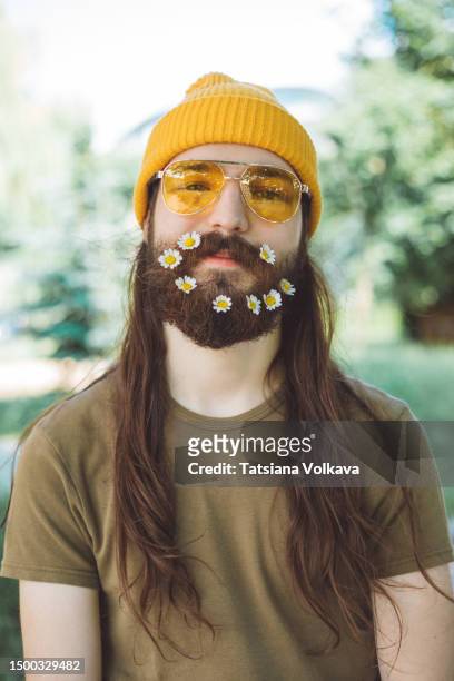 happy bearded male florist with long brown hair looking at camera with playful smirk and daisies in beard - long beard stockfoto's en -beelden