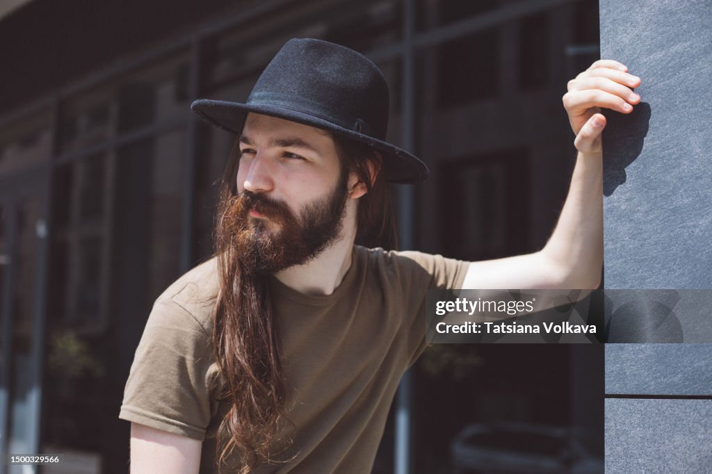 Portrait of relaxed bearded man with brown hair in black fedora standing near dark building wall